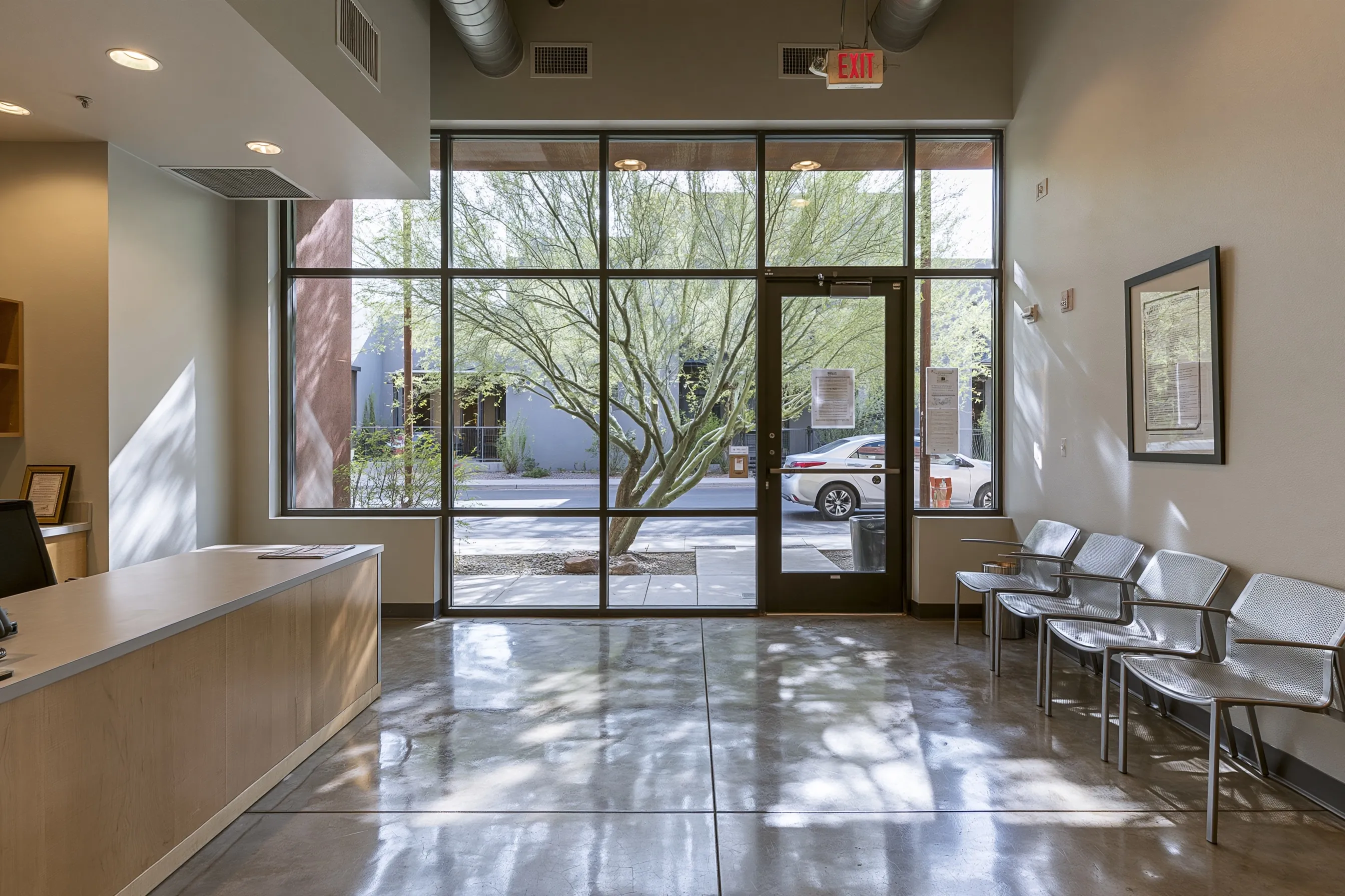 Small office reception with a polished concrete floor, a front desk, metal waiting chairs and a glass entrance with a tree outside